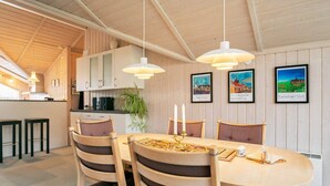 A dining area with a wooden table, chairs, and pendant lights. There are framed pictures on the wall, a plant, and a kitchen in the background. - Tranquil Oasis in Blavand-by Traum (Rudkobing)