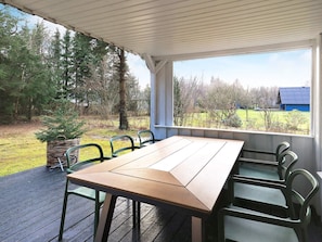 A covered outdoor area with a wooden table and chairs, a basket, and a view of a rural landscape with trees and a blue building.
