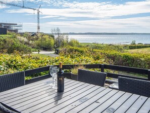 A wooden deck with a table and chairs overlooking a body of water, with a construction crane and greenery in the background.