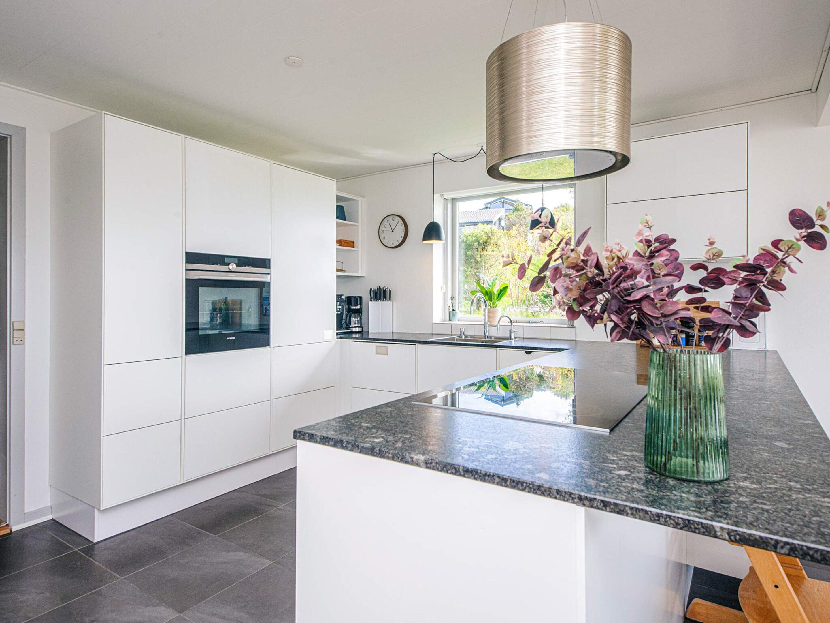 Une cuisine moderne avec des meubles blancs, un plan de travail en granit, un luminaire suspendu et une fenêtre donnant sur un paysage verdoyant.