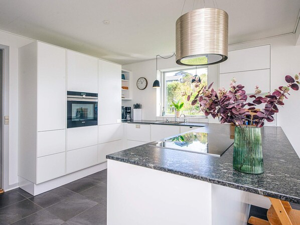 A modern kitchen with white cabinetry, a granite countertop, a hanging light fixture, and a window with a view of greenery.