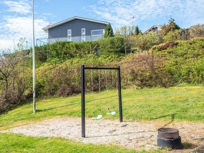 A playground with a swing set and a sandpit, surrounded by grass and bushes, with houses in the background. - 5 Star Holiday Home in Ebeltoft (Ebeltoft)