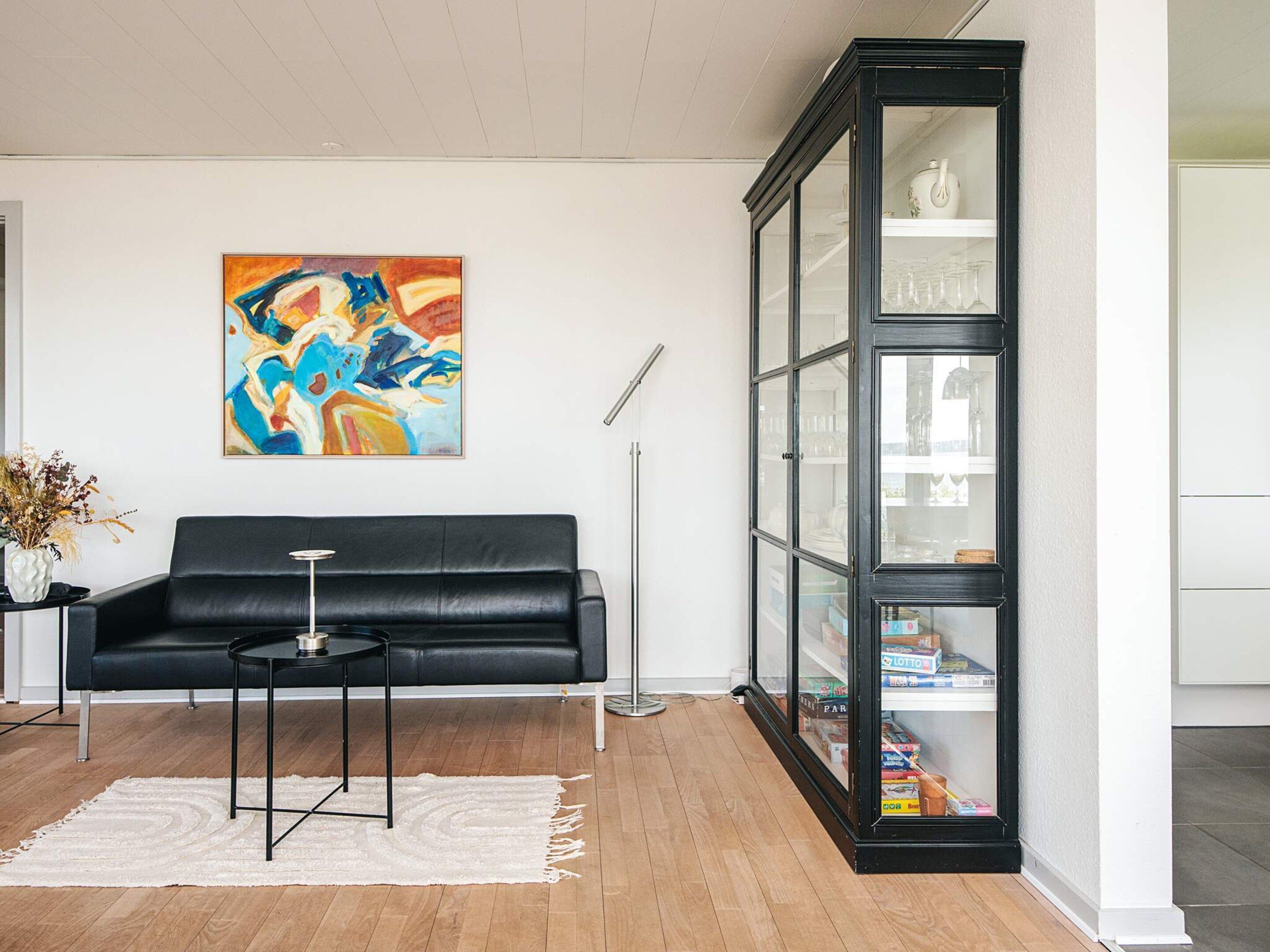 A modern living room with a black leather sofa, a small round table, a white rug, a glass cabinet with shelves, and a colorful abstract painting on the wall.