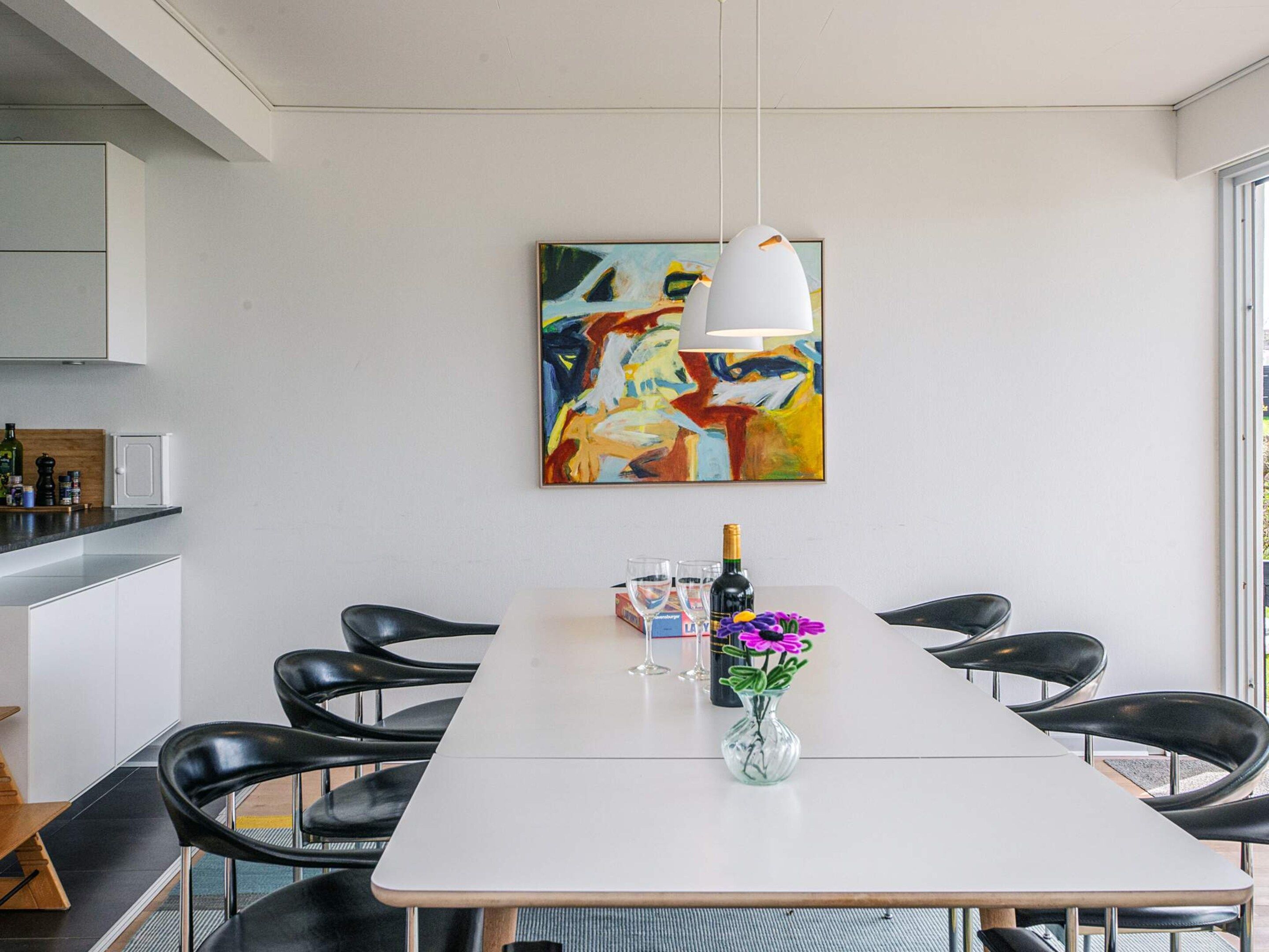 A modern dining area with a white table, black chairs, a vase with flowers, a bottle of champagne, and a colorful abstract painting on the wall.
