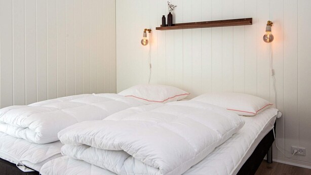 A bedroom with a bed, white bedding, a wooden shelf above the bed, and two wall-mounted lights. - 4 Star Holiday Home in Ansager (Ansager)