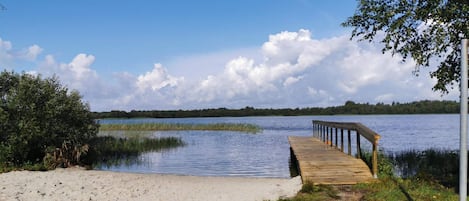 A wooden pier extending into a calm lake with a sandy shore and grassy areas.