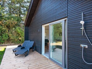 Um edifício moderno com fachada escura, porta de vidro deslizante que dá para um espaço interior e um deck de madeira com duas cadeiras de descanso.