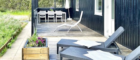 Una terraza con mobiliario exterior, una jardinera de madera con flores y un edificio de fachada oscura.