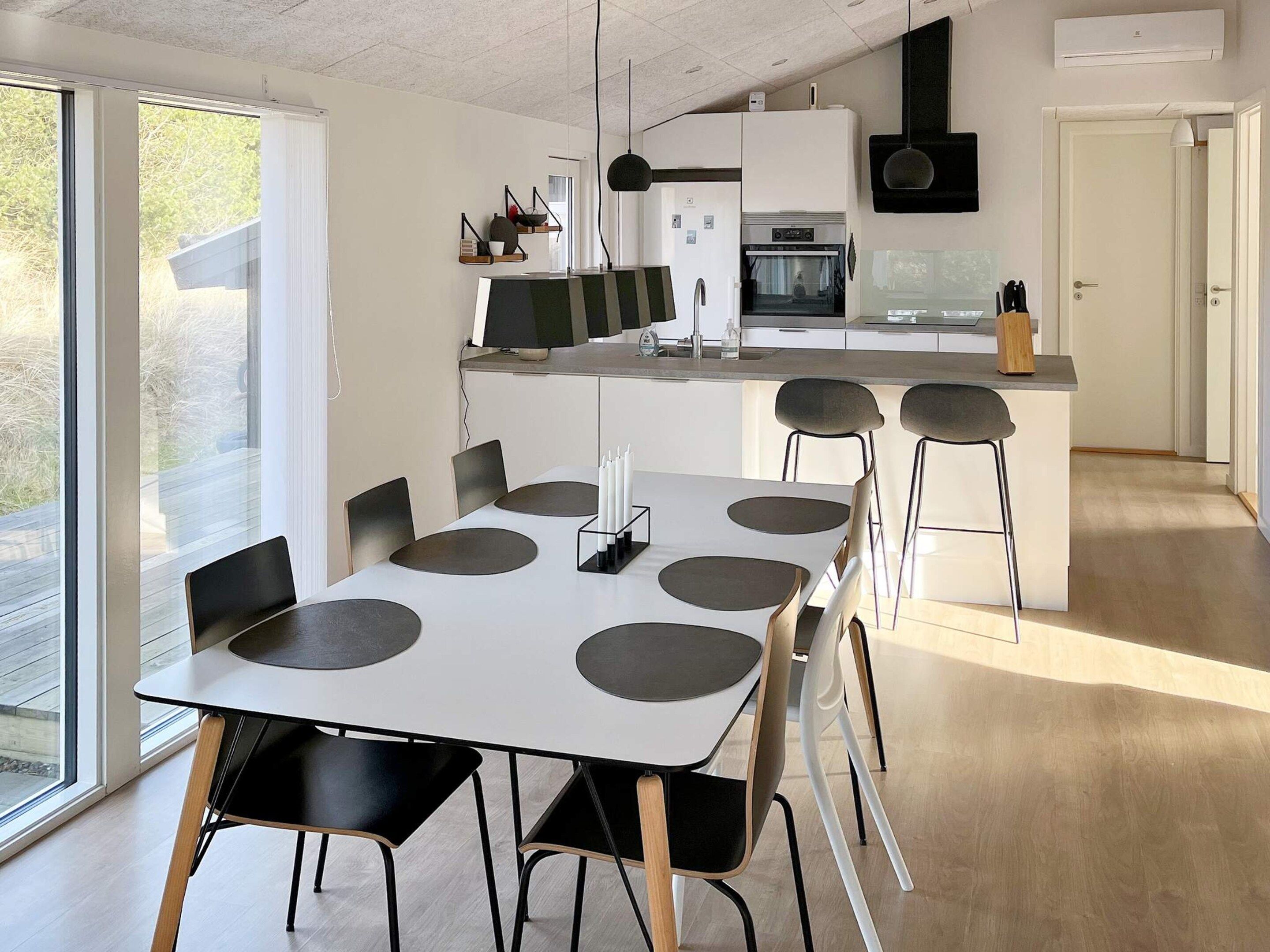 A modern kitchen with a central island, a dining area with a white table and black chairs, and a large window providing natural light.