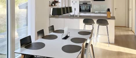 A modern kitchen with a central island, a dining area with a white table and black chairs, and a large window providing natural light.