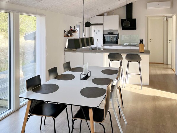 A modern kitchen with a central island, a dining area with a white table and black chairs, and a large window providing natural light.