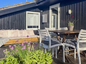 A deck with a wooden table and white chairs, a cushioned bench, and a potted plant. - 6 Person Holiday Home in Saltum (Saltum)