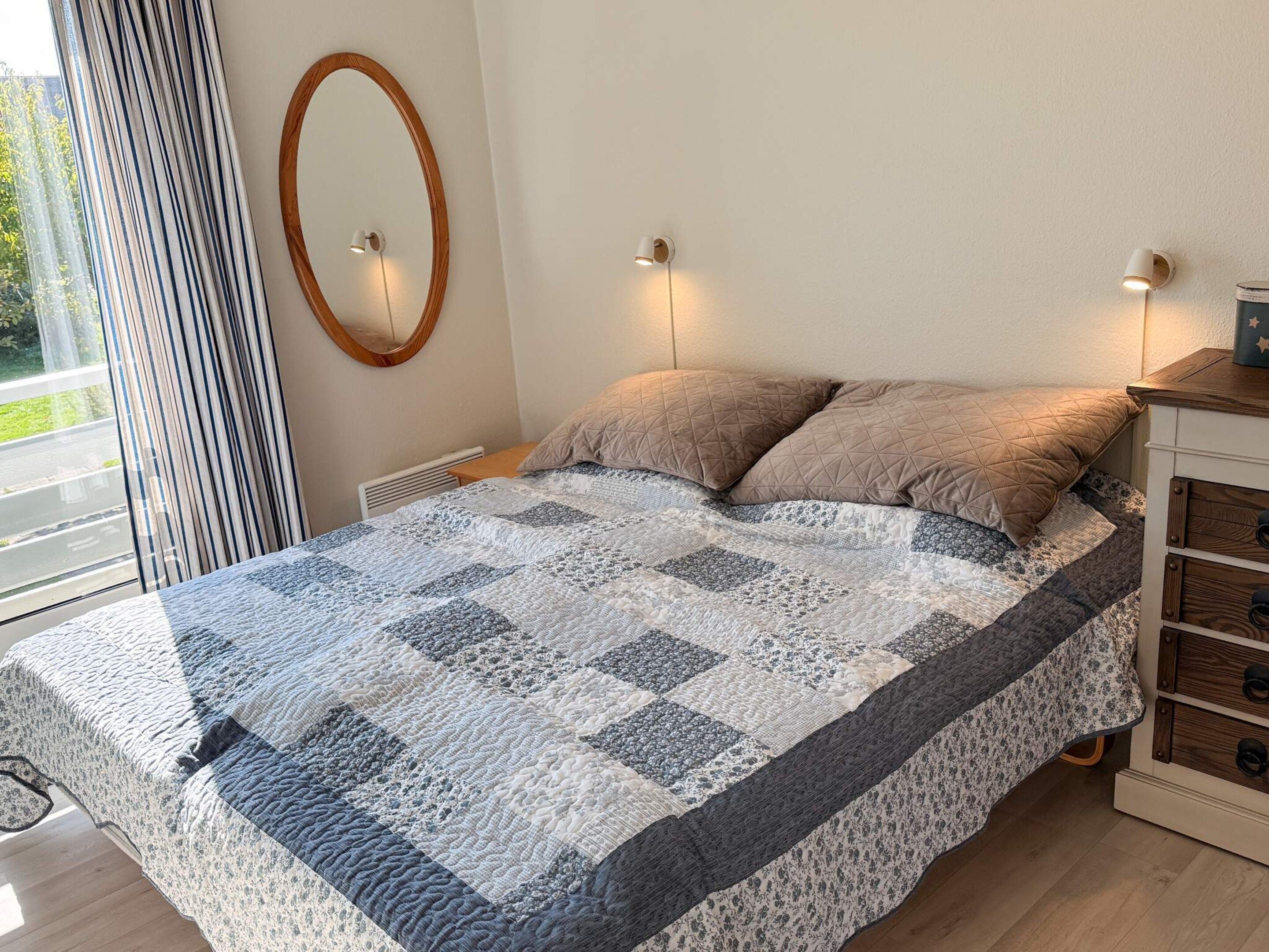 A bedroom with a bed, two bedside lamps, a wooden nightstand, a window with curtains, and a wall-mounted mirror.