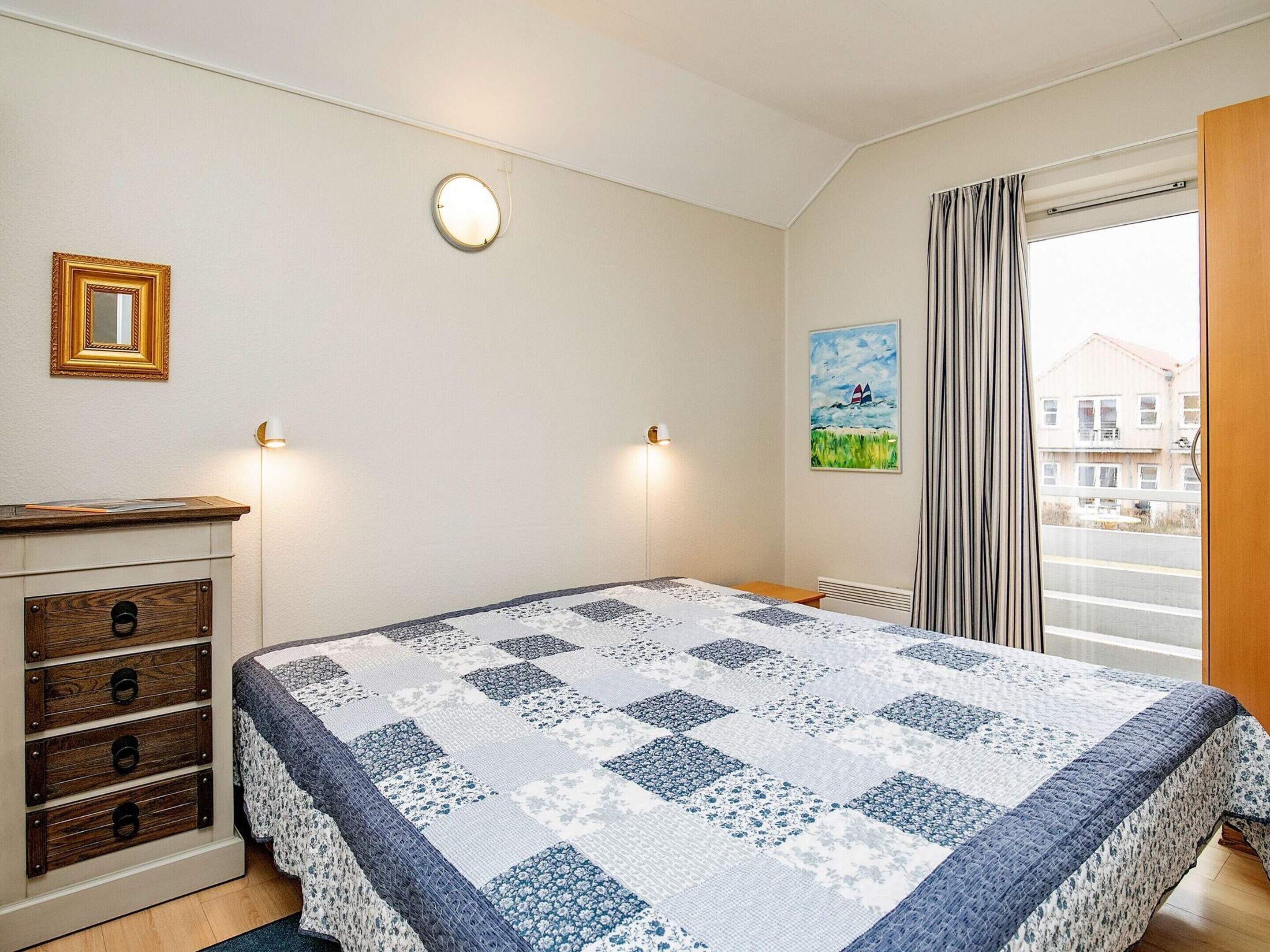 A bedroom with a bed, dresser, wall-mounted lights, a painting, and a view through a window.