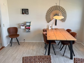 A dining area with a wooden table, four chairs, a hanging light fixture, and a wall-mounted bookcase. - 4 Person Holiday Home in Rudkobing (Rudkobing)
