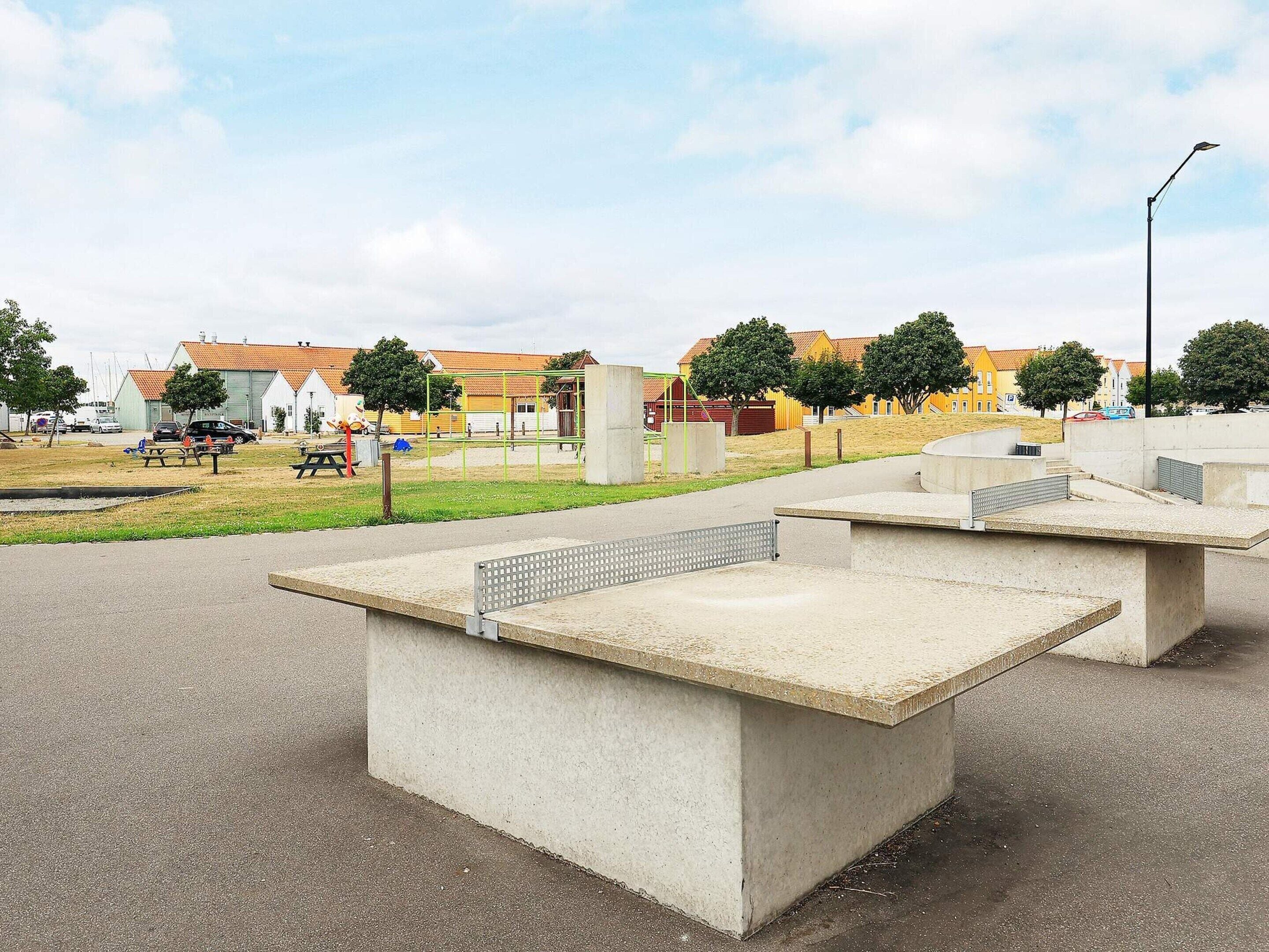 A concrete outdoor seating area with benches and tables, surrounded by a grassy field and buildings in the background.