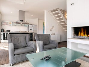 A modern living room with a glass coffee table, two grey armchairs, a fireplace, and a kitchen in the background.