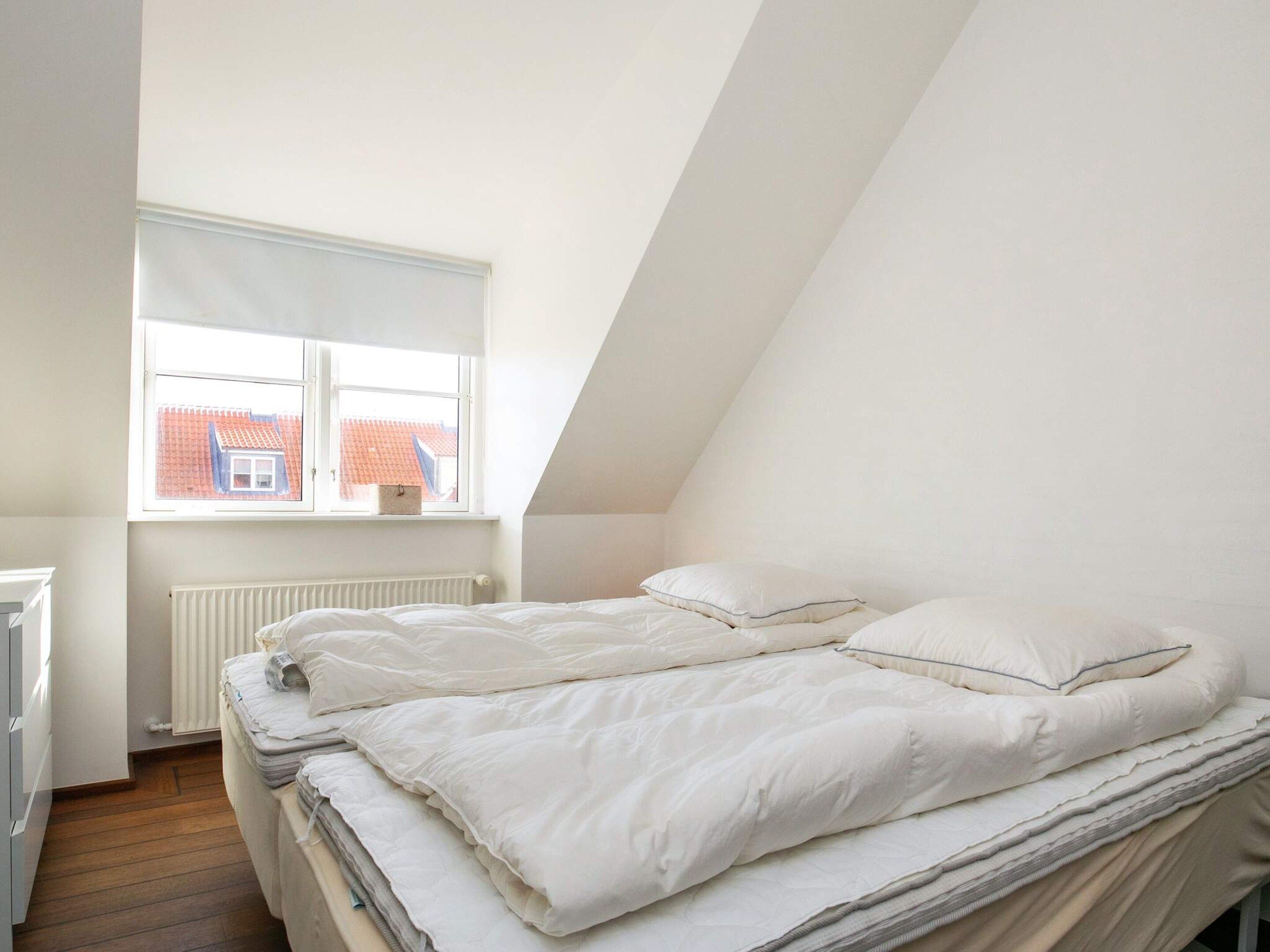 Ein Schlafzimmer mit schrägen Wänden, einem Fenster mit Blick auf ein Gebäude mit rotem Dach und einer Heizung.