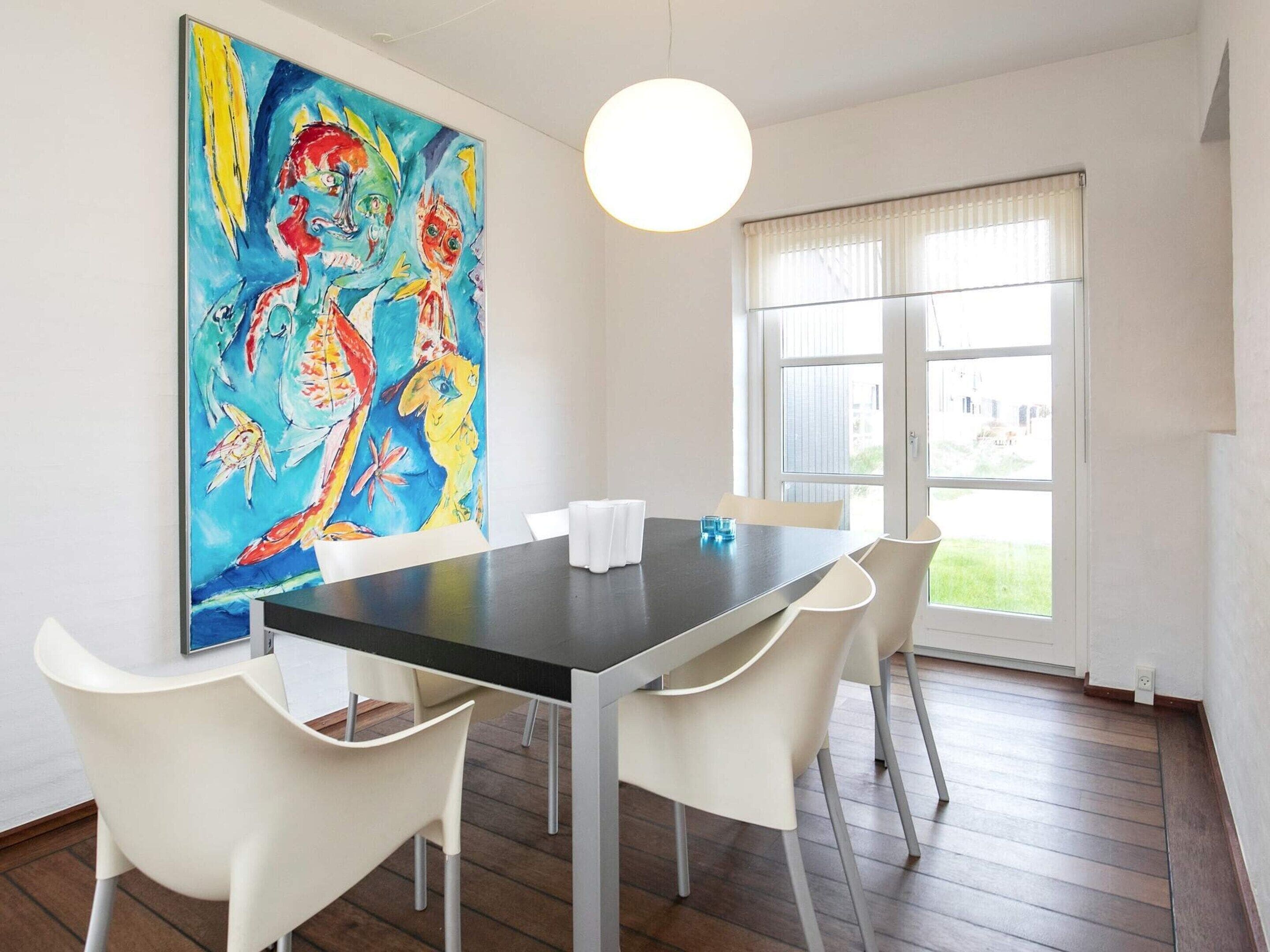 A dining area with a table and chairs, a large colorful painting on the wall, and a window with blinds.