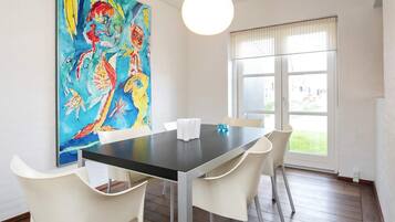 A dining area with a table and chairs, a large colorful painting on the wall, and a window with blinds.