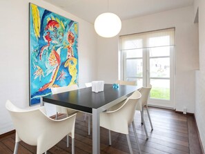 Un coin repas avec une table et des chaises, un grand tableau coloré au mur et une fenêtre avec des stores.
