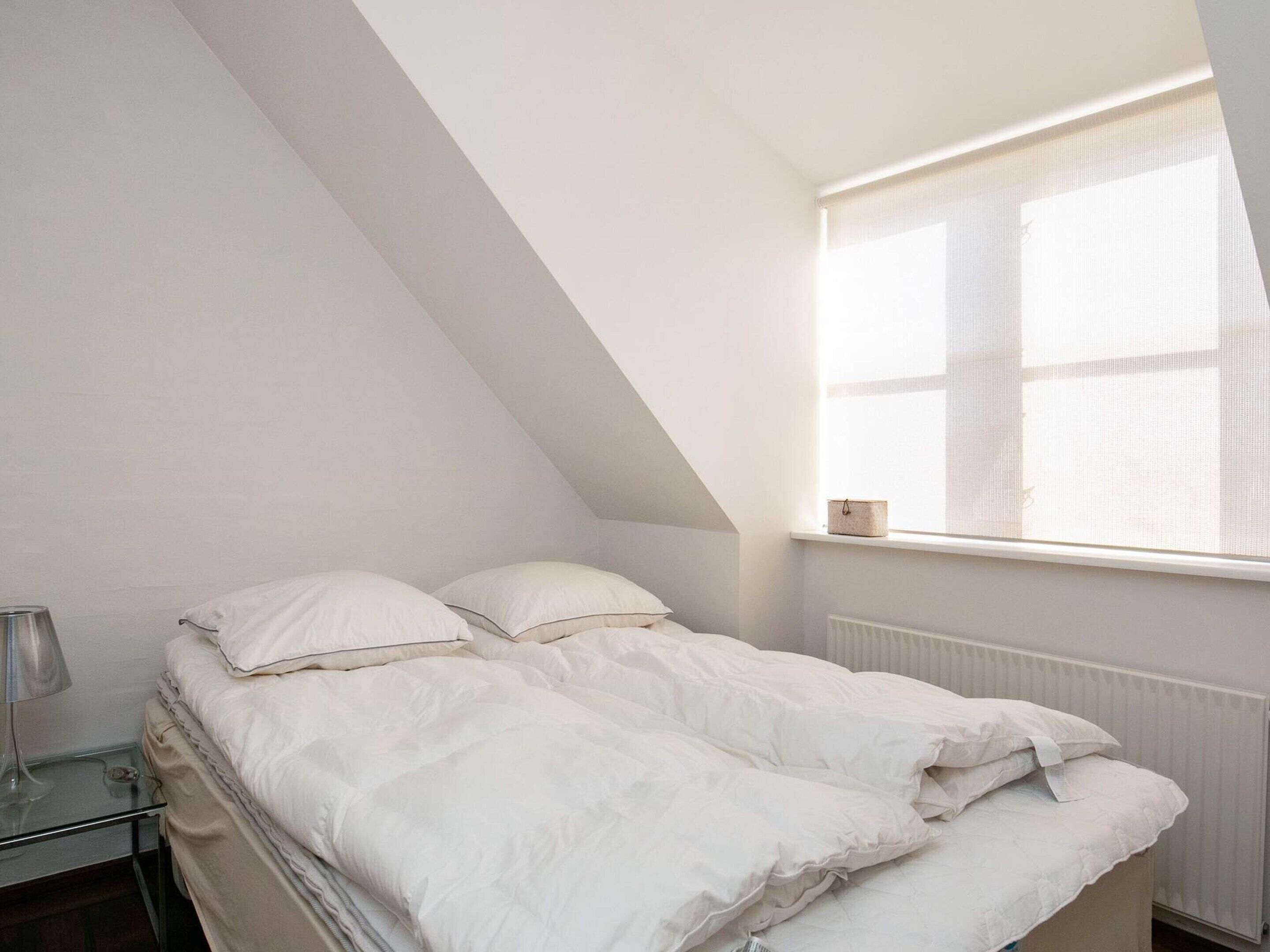 A bedroom with a sloped ceiling, a bed with white bedding, a bedside table with a lamp, and a window with a curtain.