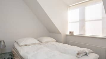A bedroom with a sloped ceiling, a bed with white bedding, a bedside table with a lamp, and a window with a curtain.