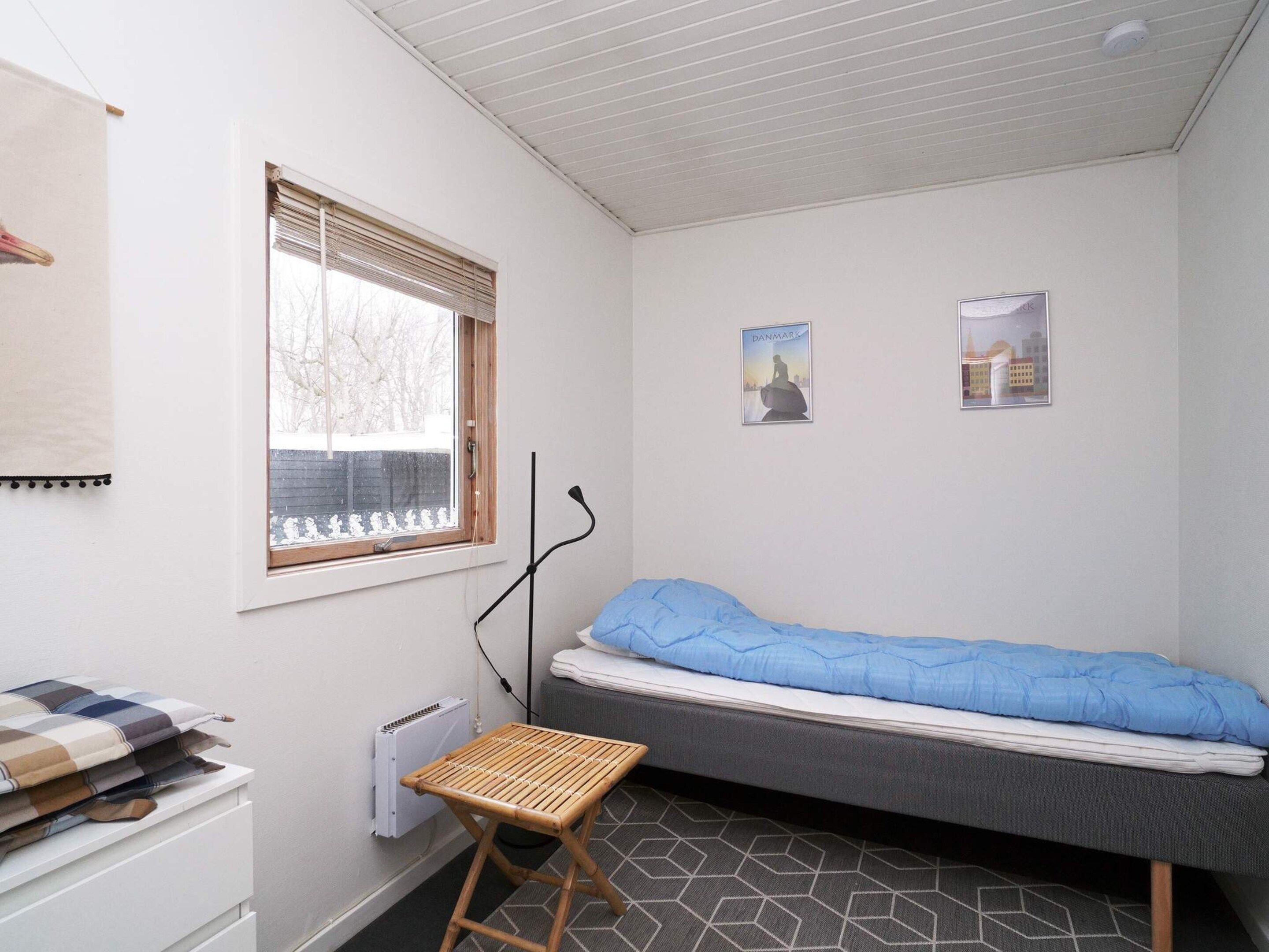 Een kleine, eenvoudig ingerichte kamer met een enkel bed, een radiator, een raam met jaloezieën en twee fotolijstjes aan de muur.