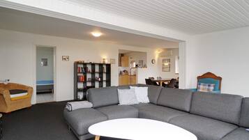 En moderne stue med en grå hjørnesofa, et hvidt sofabord, en reol og et spiseområde i baggrunden.