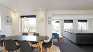 Een moderne woonkamer met een eetgedeelte, met een ronde tafel, stoelen, een bank en een groot raam met jaloezieën.