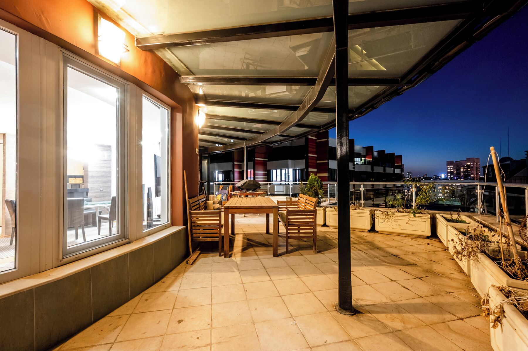 Une terrasse extérieure couverte, dotée d’un espace repas, de grandes fenêtres et offrant une vue sur les toits de la ville au crépuscule.
