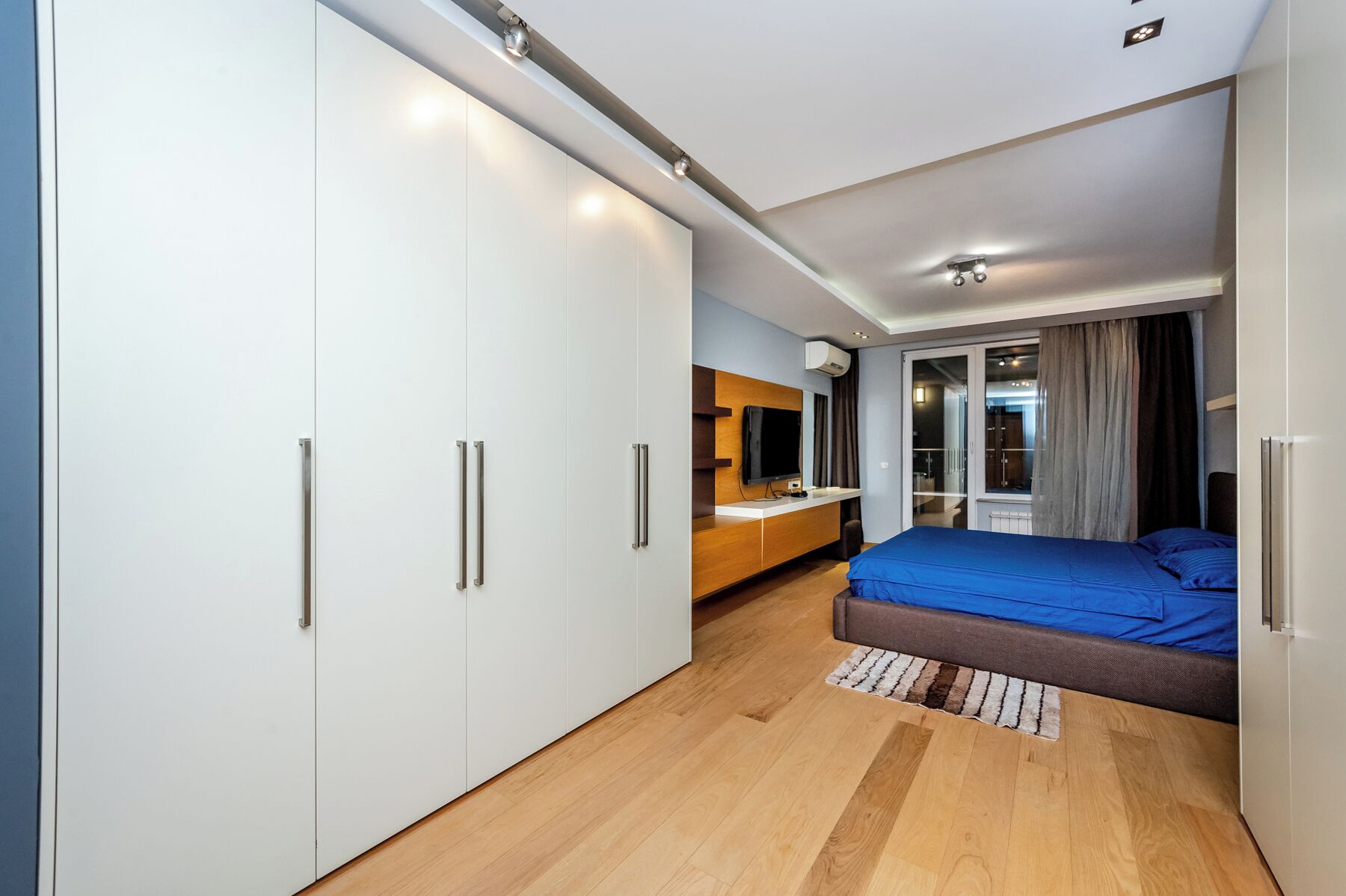 A modern bedroom with a large bed, a wardrobe, a desk, and a television.