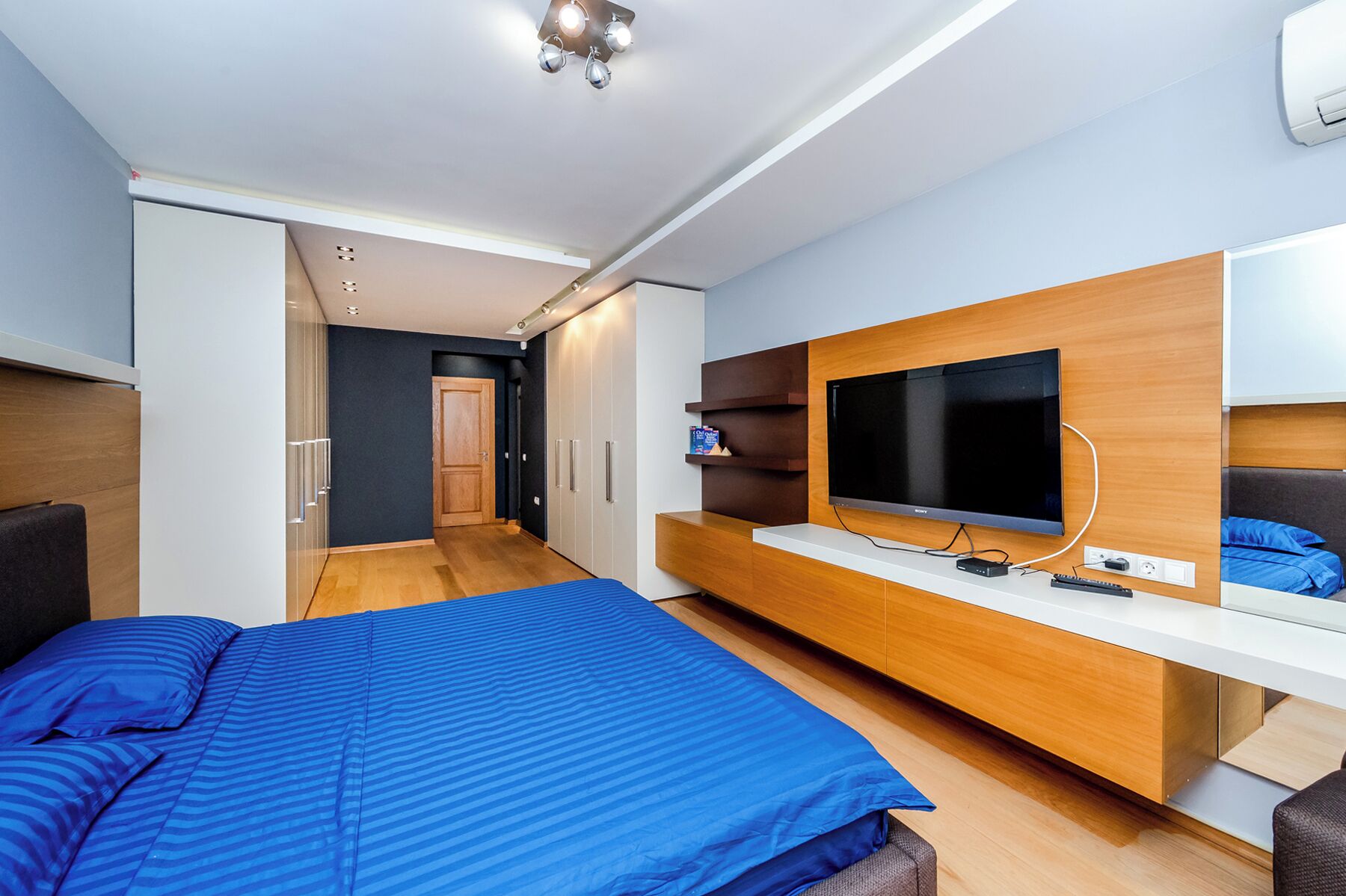 Une chambre moderne avec un grand lit, un téléviseur fixé sur une console en bois et un dressing.