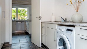 A modern kitchen with a built-in washing machine, a window with a view of greenery, and a vase with dried flowers on the countertop.