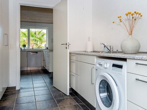 A modern kitchen with a built-in washing machine, a window with a view of greenery, and a vase with dried flowers on the countertop. - 6 Person Holiday Home in Grenaa (Grenaa)