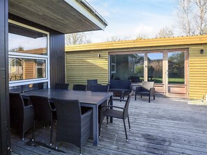 An outdoor patio with a wooden deck, a table with chairs, and a sliding glass door leading to a garden.