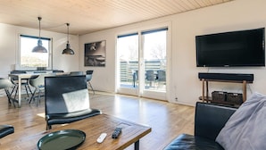 A modern living room with a wooden ceiling, a large flat-screen TV mounted on the wall, a leather sofa, and a dining area with a table and chairs. - 4 Star Holiday Home in Albaek (Aalbaek)