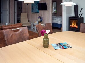 Une pièce chaleureuse avec une table à manger en bois, un vase rempli de fleurs et un magazine. On y trouve un lit, avec un feu de cheminée en arrière-plan et un téléviseur accroché au mur.