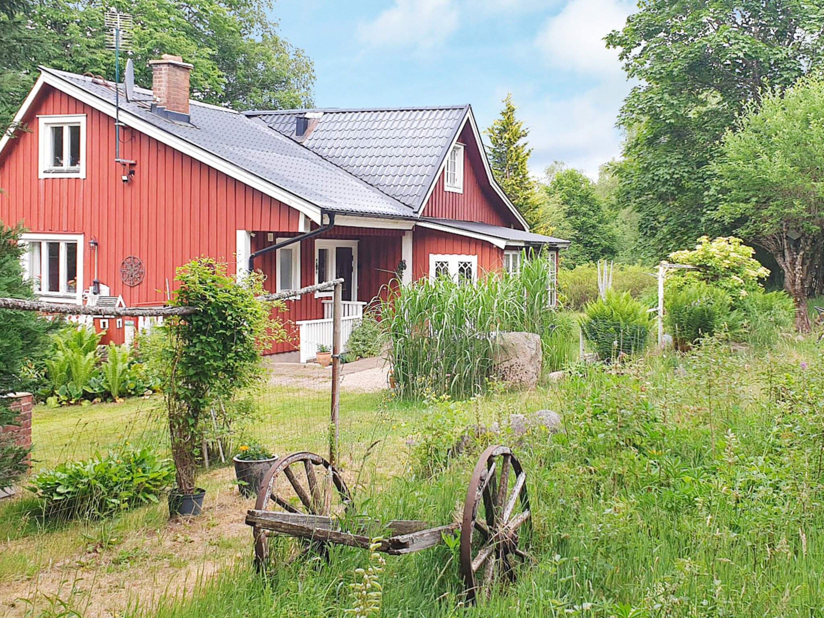 Une maison rouge avec un toit en pente, un porche et un jardin agrémenté de plantes et d’une roue en bois.