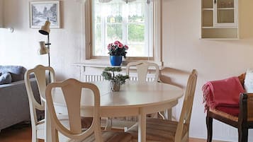 Un comedor con una mesa redonda, cuatro sillas, un florero con flores, una ventana con cortinas, una lámpara y un cuadro en la pared.