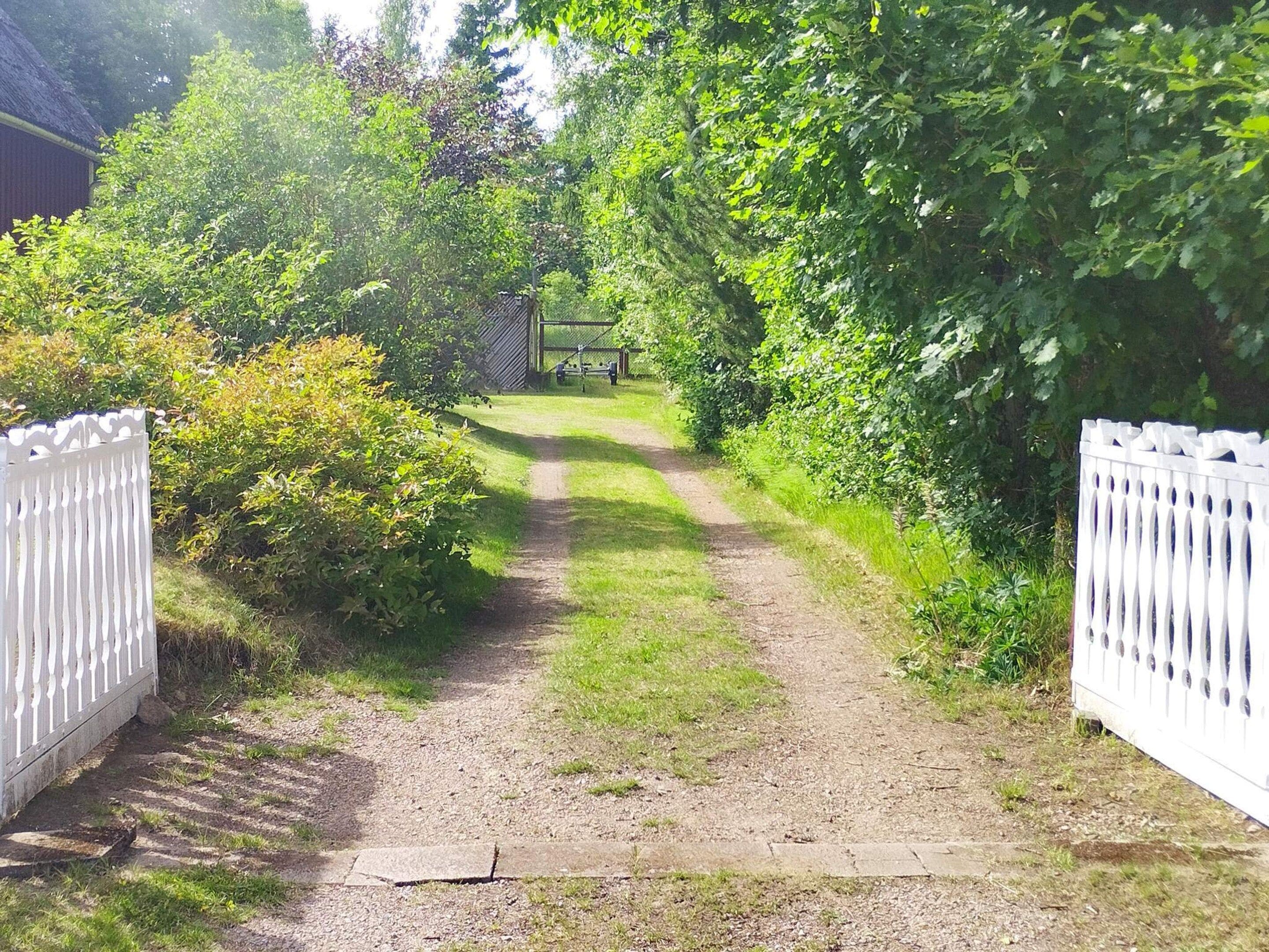 A gravel path leading to a white gate, surrounded by dense greenery and a wooden structure in the background.
