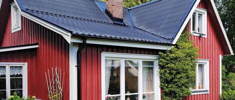 A red wooden house with a dark roof, white window frames, and a small garden area with a white fence and a tree.