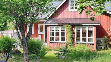 A red wooden house with white windows and a metal roof, surrounded by greenery and rocks.