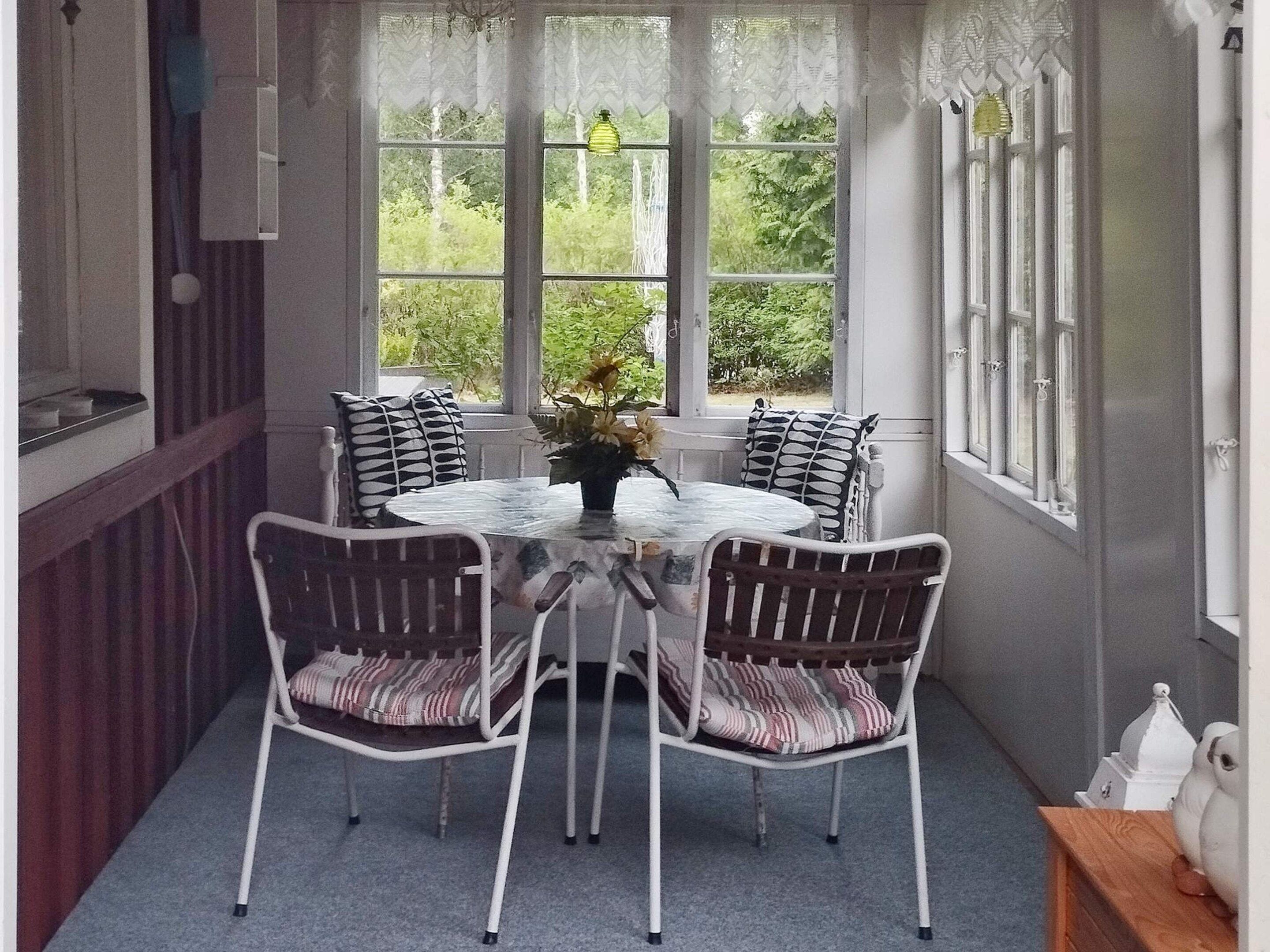 A room with a round table and four chairs, a window with lace curtains, and a view of greenery outside.