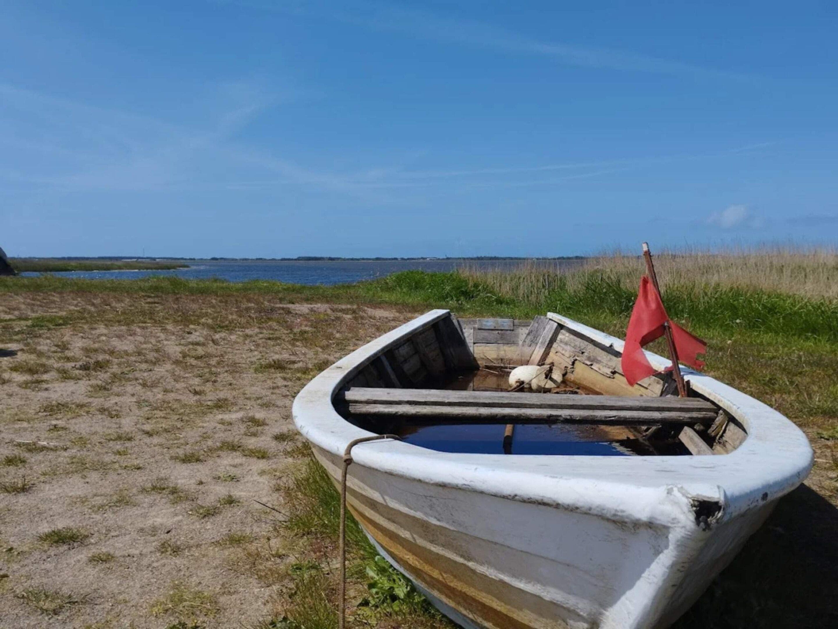 Ein kleines, weißes Ruderboot mit einer roten Flagge, teilweise im Sand versunken in der Nähe eines Gewässers.