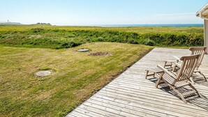 Une terrasse en bois avec des sièges extérieurs, offrant une vue sur un champ verdoyant et l’océan.