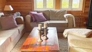 Une pièce confortable, aménagée comme un chalet, avec un canapé beige, une table basse en bois ornée de bougies et une fenêtre donnant sur la forêt.
