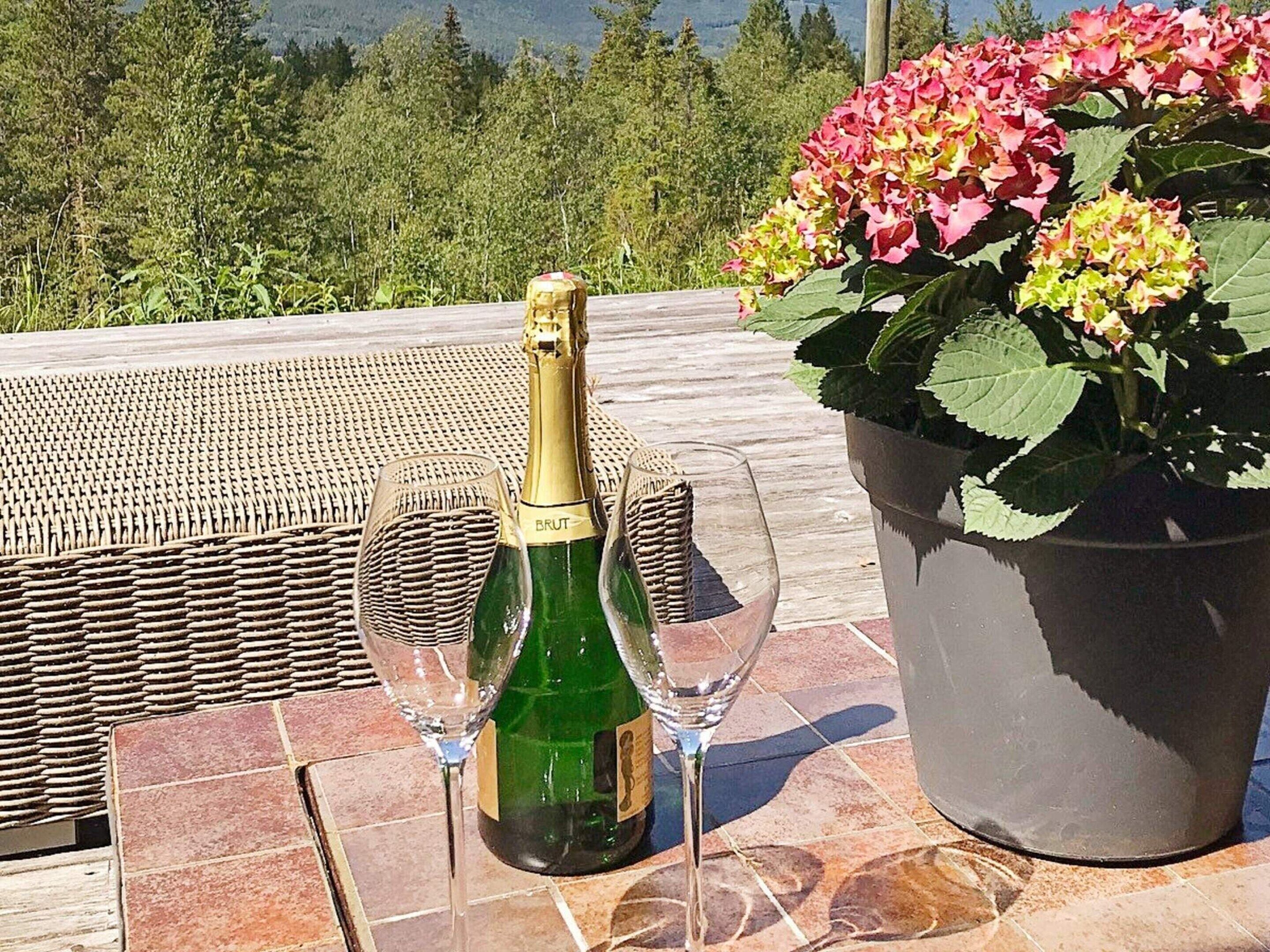 Eine Champagnerflasche und zwei Gläser auf einer gefliesten Terrasse, mit einem Korbstuhl und einem Blumentopf.