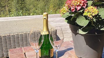 Eine Champagnerflasche und zwei Gläser auf einer gefliesten Terrasse, mit einem Korbstuhl und einem Blumentopf.