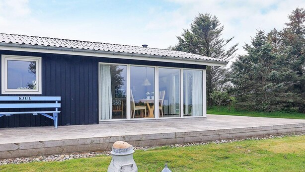 Une maison moderne à l’extérieur bleu, dotée de grandes portes vitrées, d’une terrasse en bois et d’un banc portant l’inscription « KJUL ».